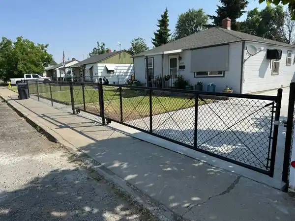 A black chain-link rolling gate across a residential driveway, fence continuing along the sidewalk
