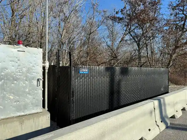 Black slatted chain link utility enclosure behind a concrete barrier