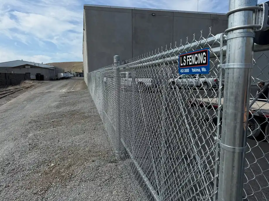 A galvanized commercial chain-link fence by L S Fencing beside a concrete building, the blue L S Fencing sign hanging on the fabric