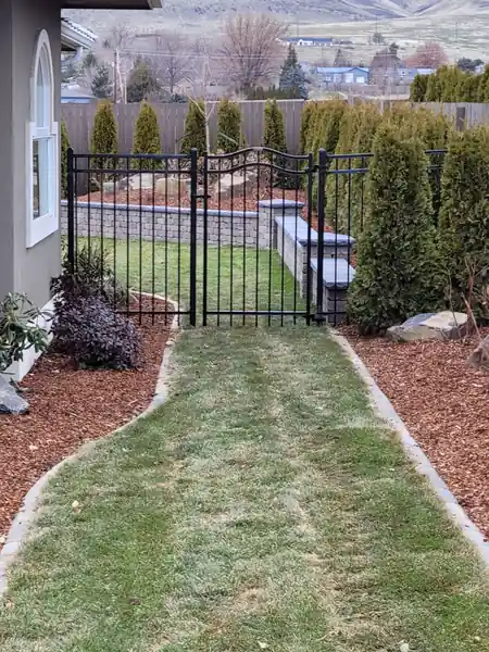 A black ornamental gate and fence across a sod path, Yakima hills behind