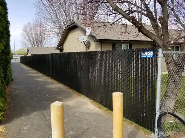 Black slat privacy chain link running along a paved path with the blue L S Fencing sign hanging on it