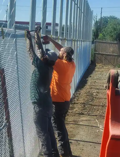 Two L S Fencing crew members tying chain-link fabric to a tall fence