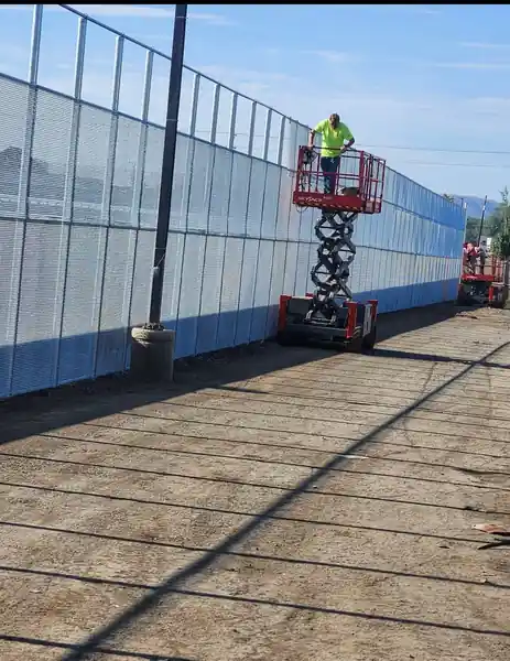 An L S Fencing crew member on a scissor lift stringing a tall screen onto a chain-link fence